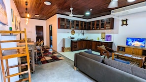 A living room with a sofa, a wooden ladder, a kitchen with wooden cabinets, a television, and a ceiling fan. - Kay's Place Train House Jambo (Zanzibar Town)