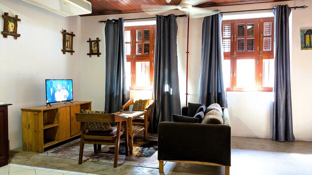 A living room with a television, wooden furniture, a sofa, and large windows with curtains. - Kay's Place Train House Jambo (Zanzibar Town)