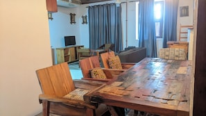 A dining area with a long wooden table, several chairs, a sofa, and a television in the background. - Kay's Place Train House Jambo (Zanzibar Town)