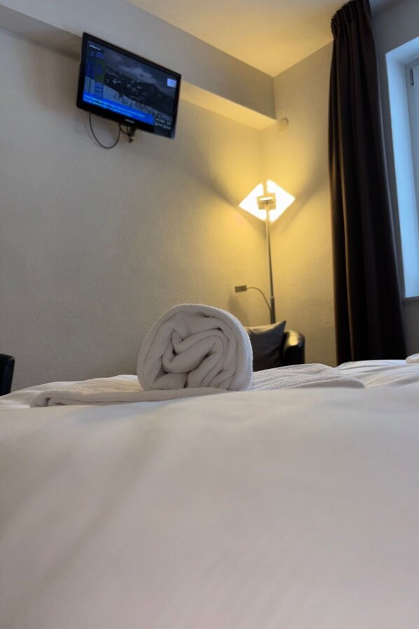 Une chambre d’hôtel avec un lit, un téléviseur fixé au mur, une lampe et une fenêtre avec des rideaux.
