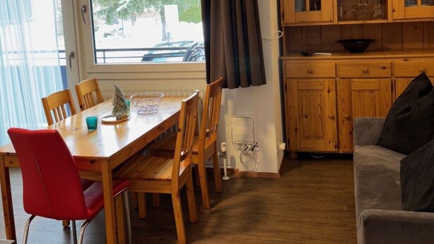A room with a wooden dining table, chairs, a television mounted on the wall, a sofa, and wooden cabinets. - 2_9 Cosy Kaprun Apartment by Interhome (Kaprun)