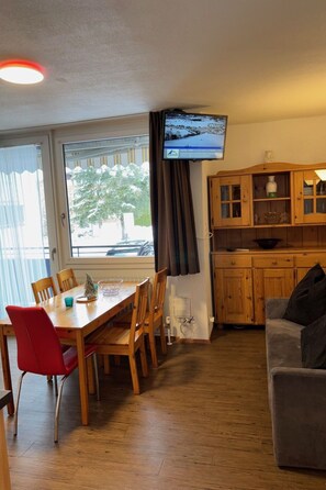 Une pièce comprenant une table à manger en bois, des chaises, un téléviseur fixé au mur, un canapé et des meubles de rangement en bois.