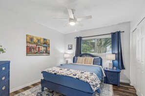 Ein Schlafzimmer mit Bett, Nachttisch, Deckenventilator, Fenster mit Vorhängen und einem Bild an der Wand.