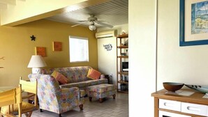 En stue med sofa, spisebord med stole, ventilator i loftet og et køkkenområde med skabe og komfur.