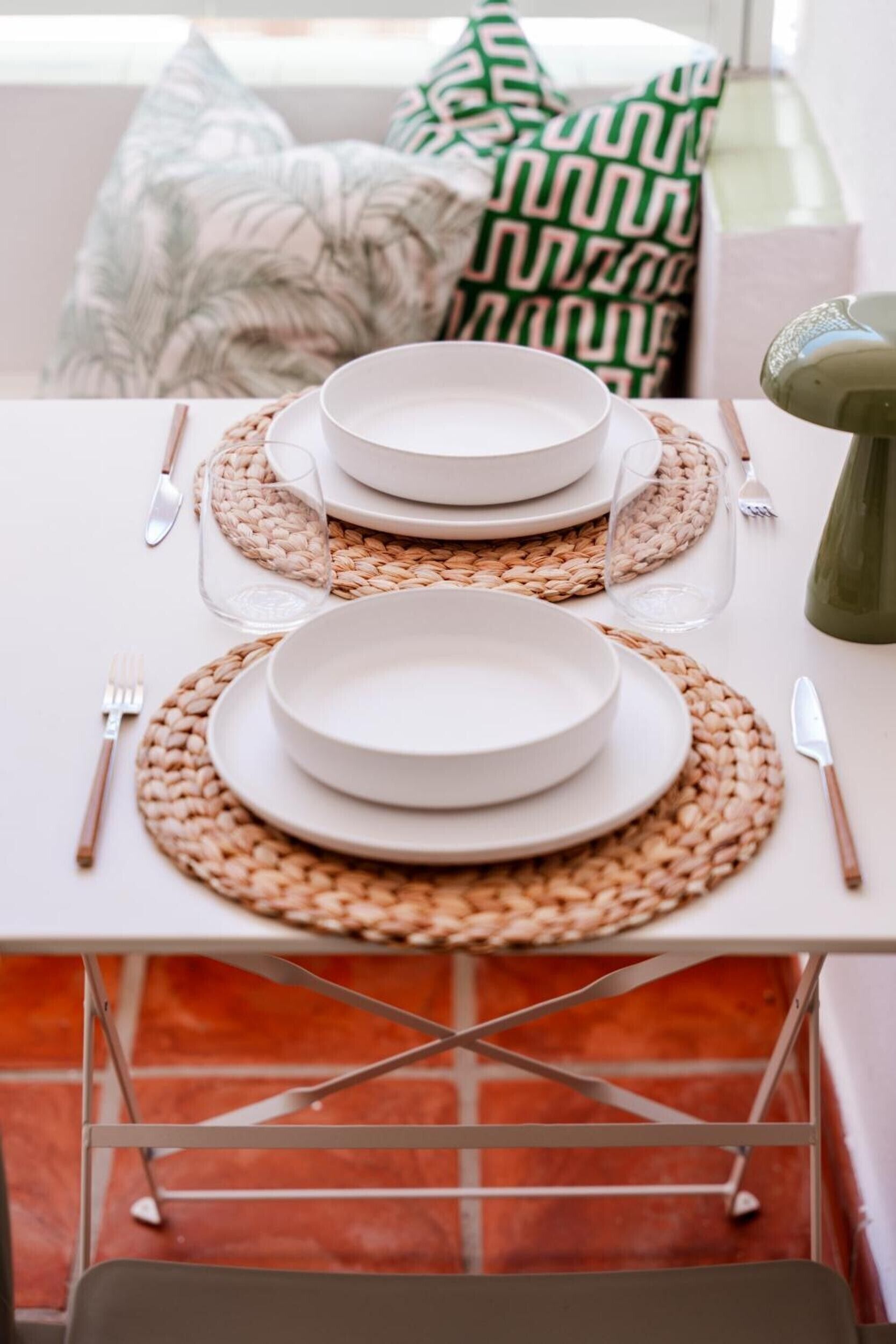 A dining table set with white plates, woven placemats, and cutlery, with a green bottle and a patterned pillow in the background.