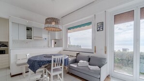 Una cuina moderna amb armaris blancs, una taula de dinar amb estovell blau, un sofà gris i una finestra gran amb vistes.