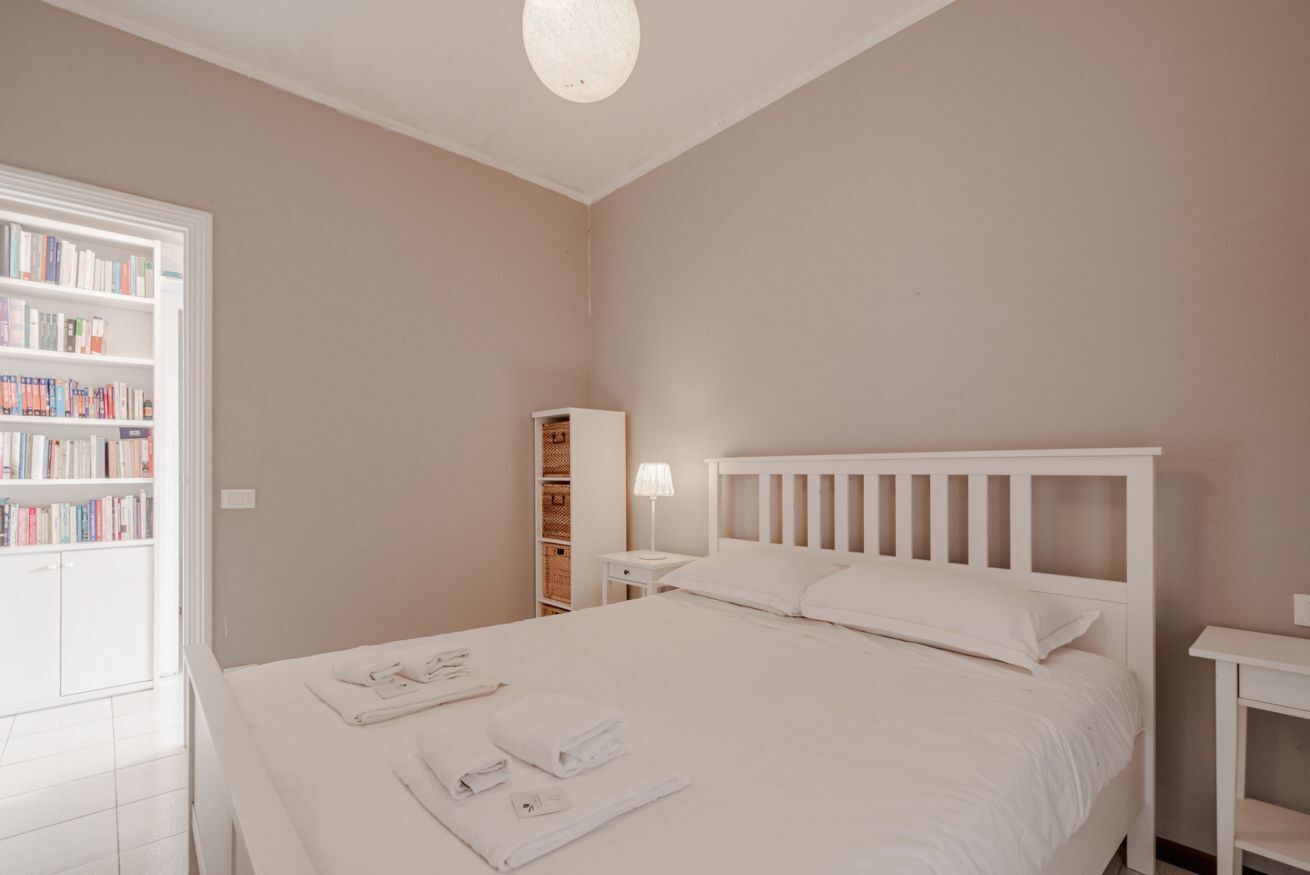A bedroom with a bed, a bedside table, a lamp, a bookshelf, and a wardrobe.