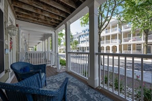 A covered porch with wicker chairs, a patterned rug, and a view of a street with multi-story buildings.