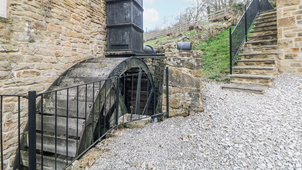 A stone building with a water wheel, gravel ground, and a metal gate. - Little Mill (Chesterfield)