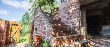 Una escalera de piedra rústica que conduce a una puerta de madera, rodeada de follaje y un cielo despejado.