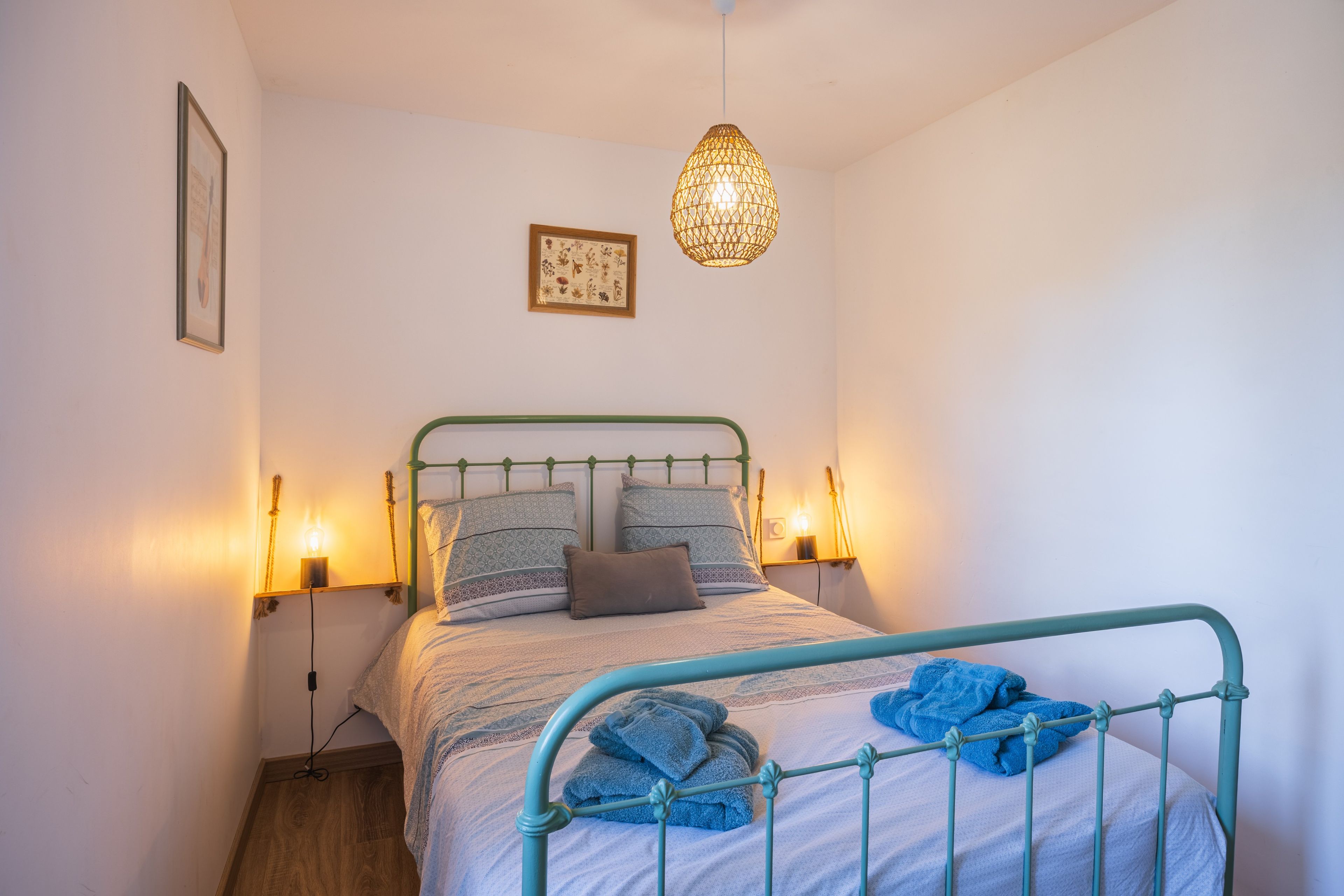 A neatly made bed with a green metal frame, a decorative hanging light, and a framed botanical print on the wall.