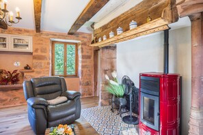A cozy room with a leather armchair, a red wood-burning stove, and a window with a view of greenery. - Ségonzac Cottage: Lake View, Private Terrace, Wi-Fi (Villecomtal)