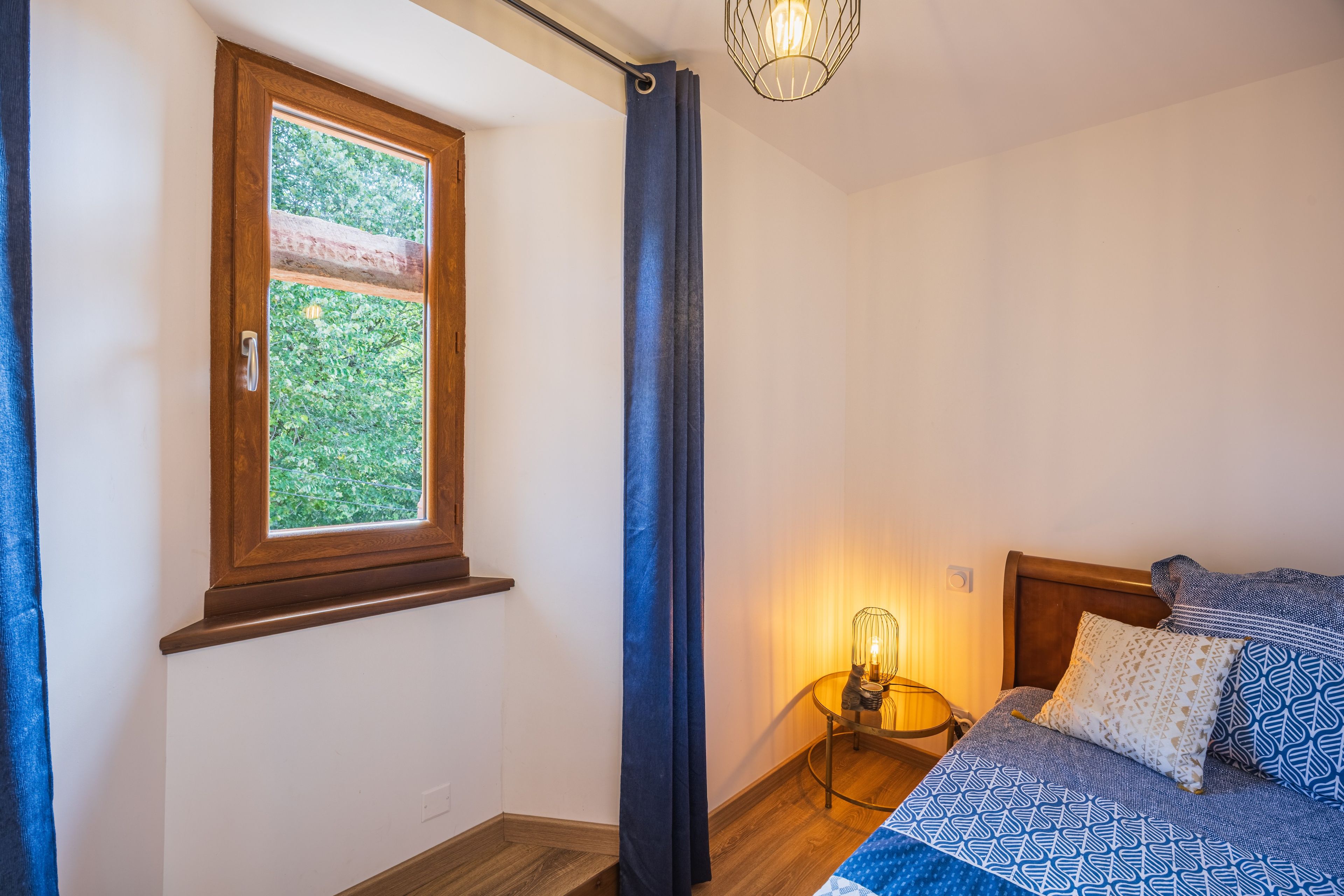 A bedroom with a wooden window, a bed with patterned bedding, a bedside table with a lamp, and a curtain.