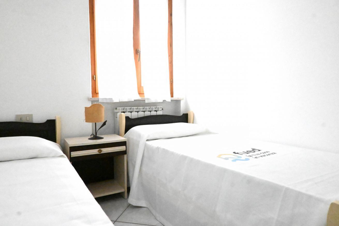 A hotel room with two beds, a nightstand, a lamp, and a window with curtains.