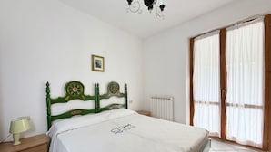 Una camera da letto con testiera verde, un letto con copripiumino bianco, un comodino con lampada, un termosifone e una finestra con tende.