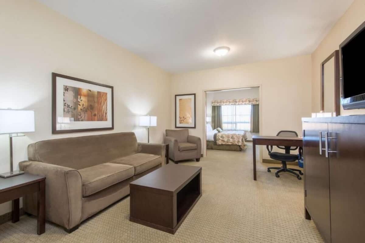Quarto de hotel com sofá, poltrona, escrivaninha e televisão.