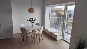 A modern kitchen with a round dining table, white chairs, a hanging light fixture, and a large window with a view of the outside. - Sea View Apartment with balcony 2BR (Argir)