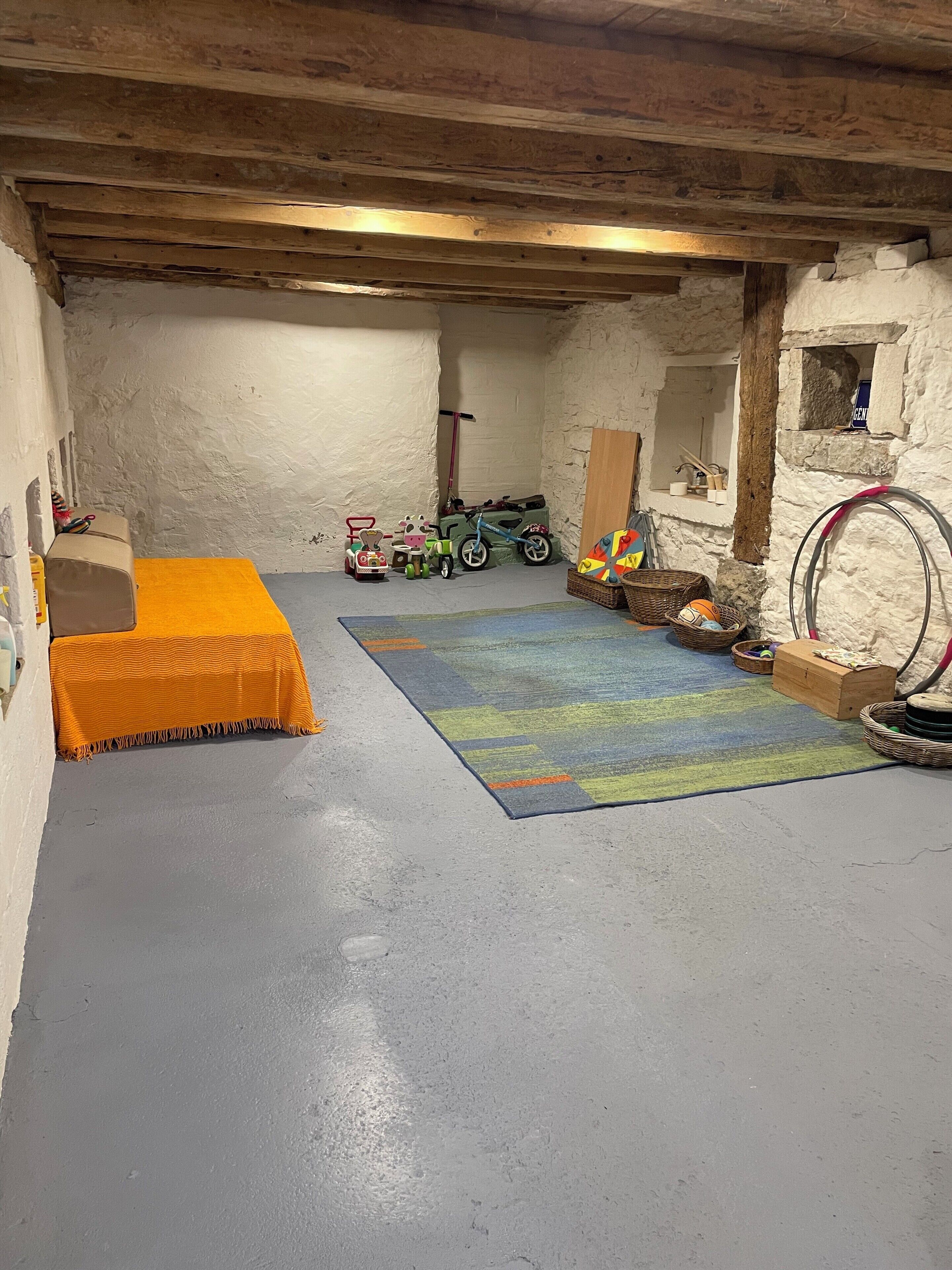 Una habitación con piso gris, una alfombra colorida, una cama con colcha naranja, un coche de juguete, un patinete y un aro de hula.