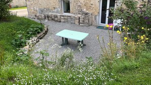 En byggnad av sten med en uteplats av grus, en ljusblå bänk och en trädgård med olika sorters blommor.