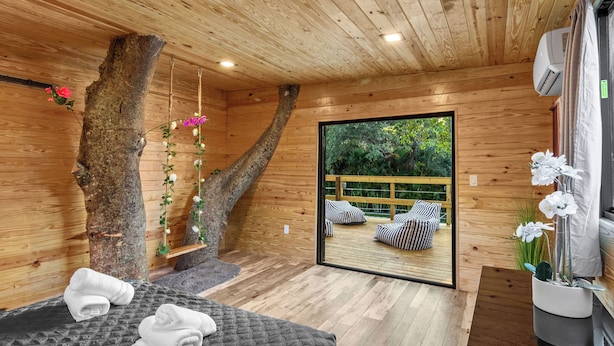 A wooden cabin interior with a bed, towels, a hanging plant, and a view of an outdoor deck.