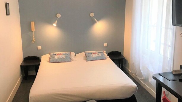 A hotel room with a bed, two bedside lamps, a nightstand, a TV, and a window with curtains.
