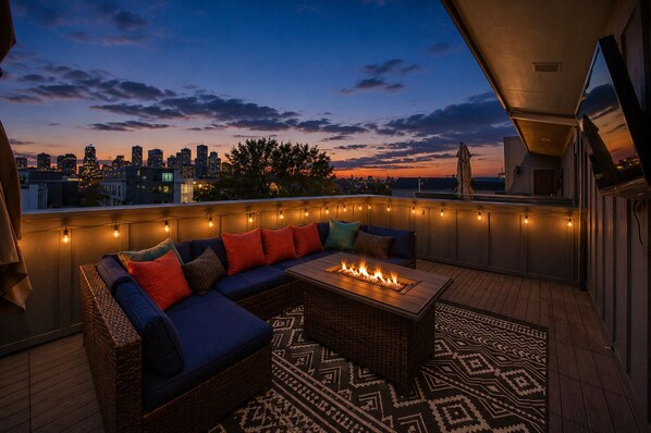 Terraza en la azotea con sofá, chimenea, luces colgantes y vistas a la ciudad.