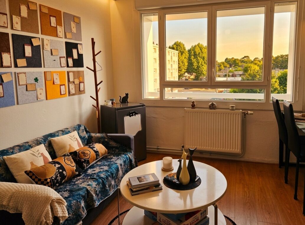 Ein Zimmer mit einer Couch, einem Tisch mit Büchern und einer Vase, einem Heizkörper und einem Fenster mit Blick auf Gebäude und Bäume.