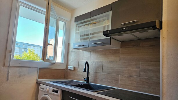Une cuisine compacte équipée d’un lave-linge, d’une cuisinière et d’une fenêtre donnant sur des immeubles.