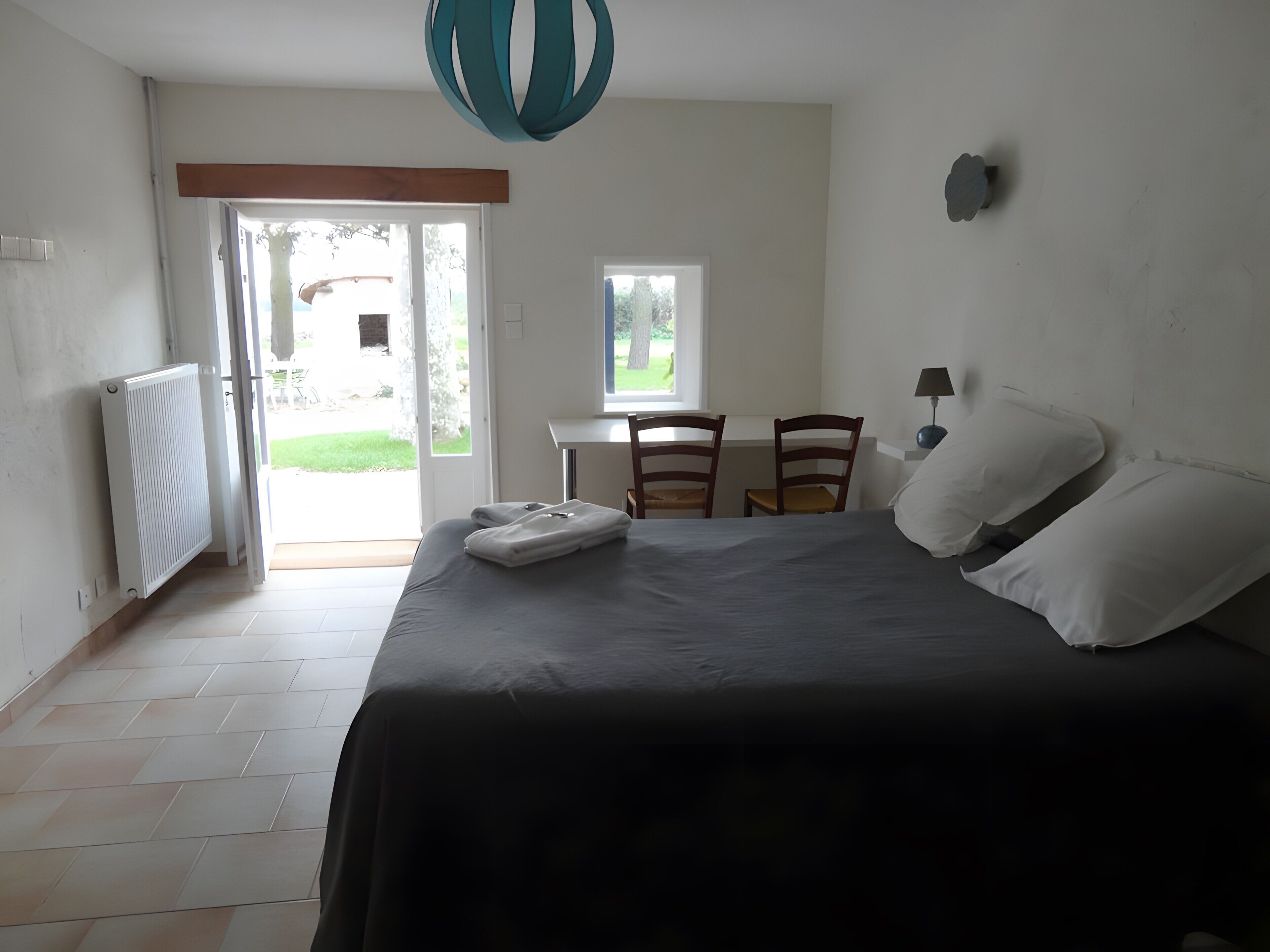 Une chambre à coucher avec un lit, deux chaises, une table, une lampe et une vue sur l’extérieur, visible à travers une porte ouverte.