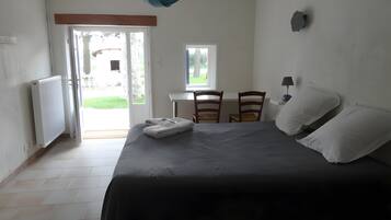 Une chambre à coucher avec un lit, deux chaises, une table, une lampe et une vue sur l’extérieur, visible à travers une porte ouverte.