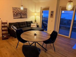 A modern living room with a sectional sofa, a round wooden dining table, and two chairs. There is a ladder, a potted plant, and a painting on the wall.