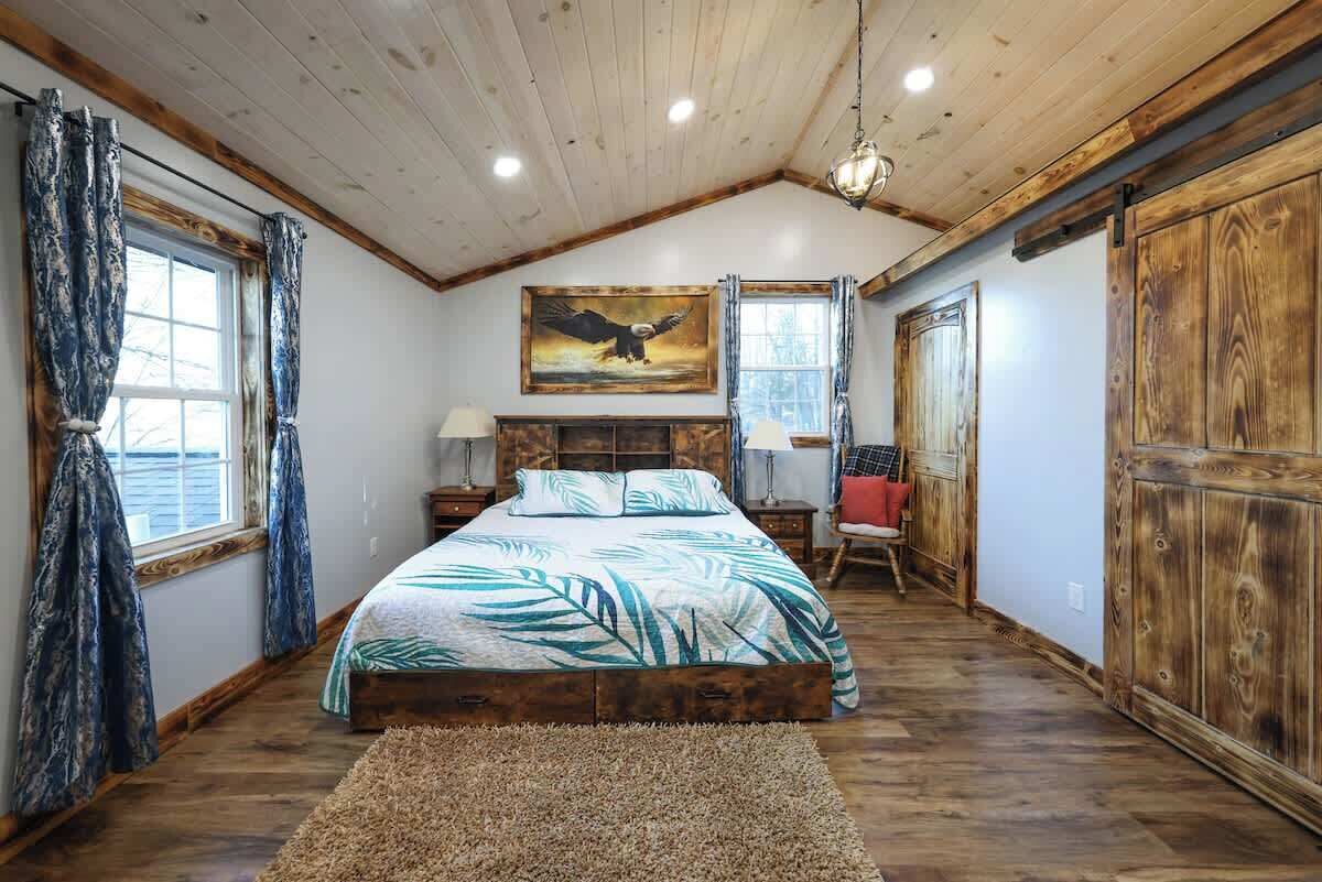 A bedroom with a wooden ceiling, a bed with a tropical-themed bedspread, a painting of an eagle, and a large wooden wardrobe.