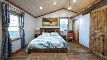 A bedroom with a wooden ceiling, a bed with a tropical-themed bedspread, a painting of an eagle, and a large wooden wardrobe.