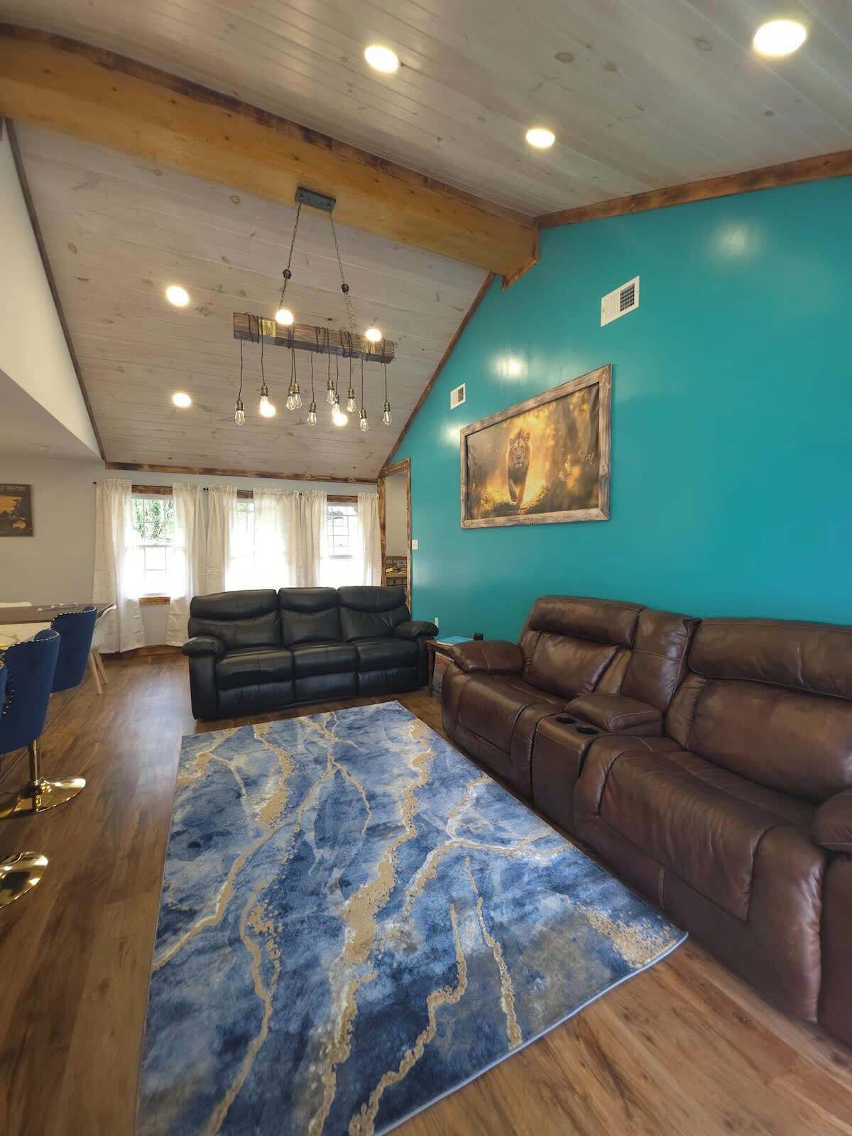 A living room with a blue wall, a brown leather sofa, a blue patterned rug, and a ceiling with wooden beams and recessed lighting.