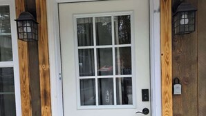 Uma porta branca com painel de vidro, ladeada por duas luminárias estilo lanterna, inserida em um moldura de madeira com parede texturizada.