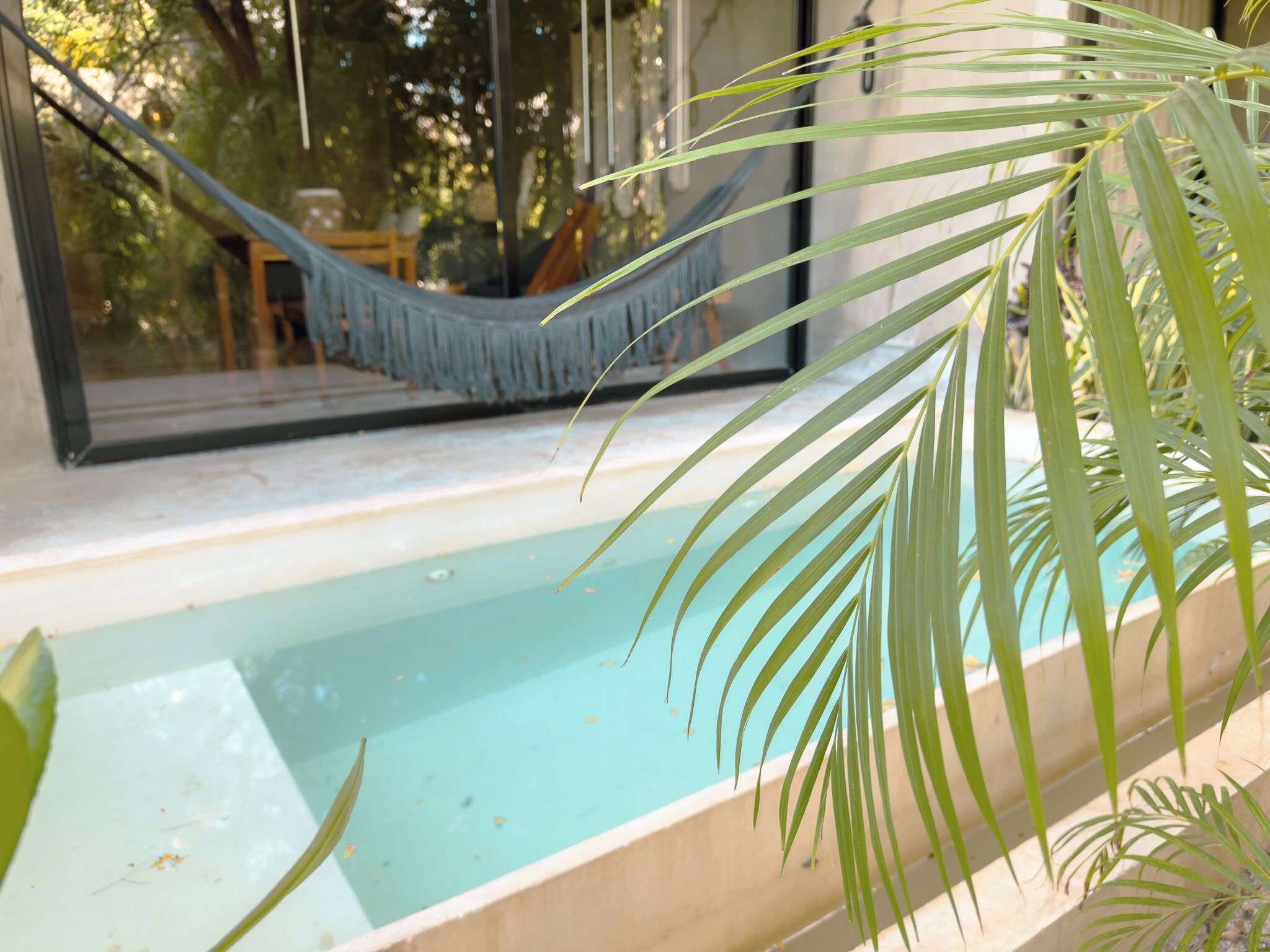 A bathtub with a view of a hammock and a window overlooking a room with a table and chairs.