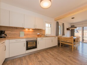 A modern kitchen with white cabinets, a built-in oven, and a dining area with a wooden table and chairs. - 1_3 Chalet Edelweiss by Interhome (Zell am See)