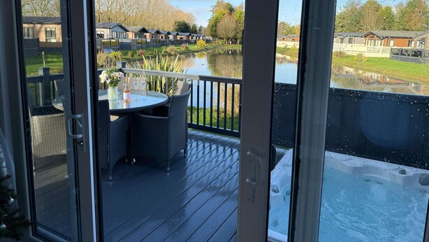 A deck with a table and chairs, a hot tub, and a view of a lake with buildings in the background. - Host & Stay | The White Swan (Allerthorpe)