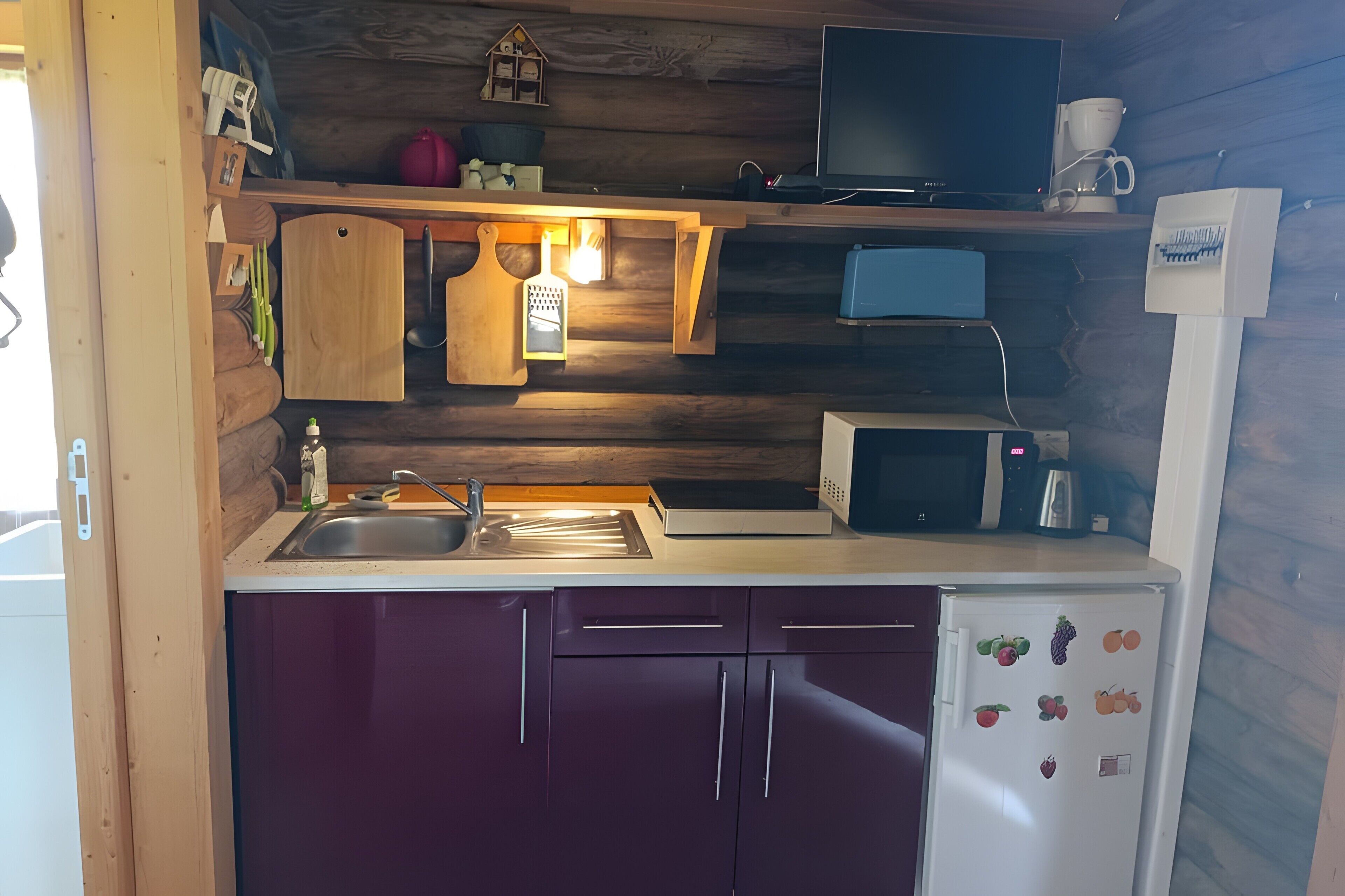 Une petite cuisine équipée d’un évier, d’un four à micro-ondes et d’un réfrigérateur, avec des murs en bois et une petite étagère où sont rangés divers objets.