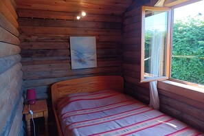 A log cabin room with a bed, a small table with a lamp, a painting on the wall, and a window with a view of greenery. - Ideal Chalet for Hiking (Valorbiquet)