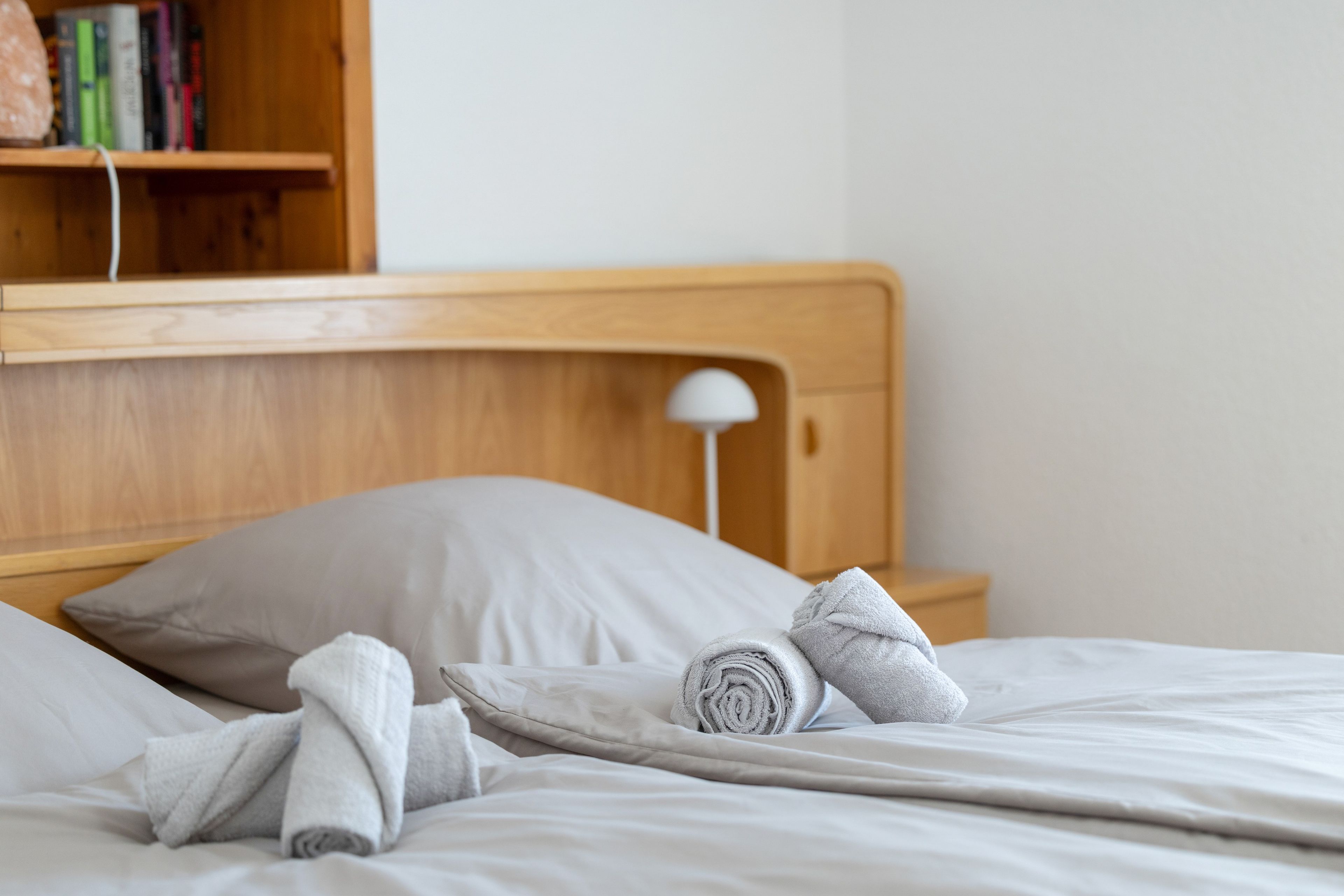 Ein ordentlich bezogenes Bett mit grauem Kopfkissen und zusammengerollten Handtüchern, ein Holz-Kopfbrett und eine Nachttischlampe.