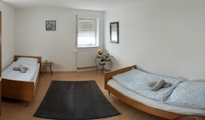 A bedroom with two beds, a radiator, a window with blinds, a small table with a vase of flowers, and a black rug.