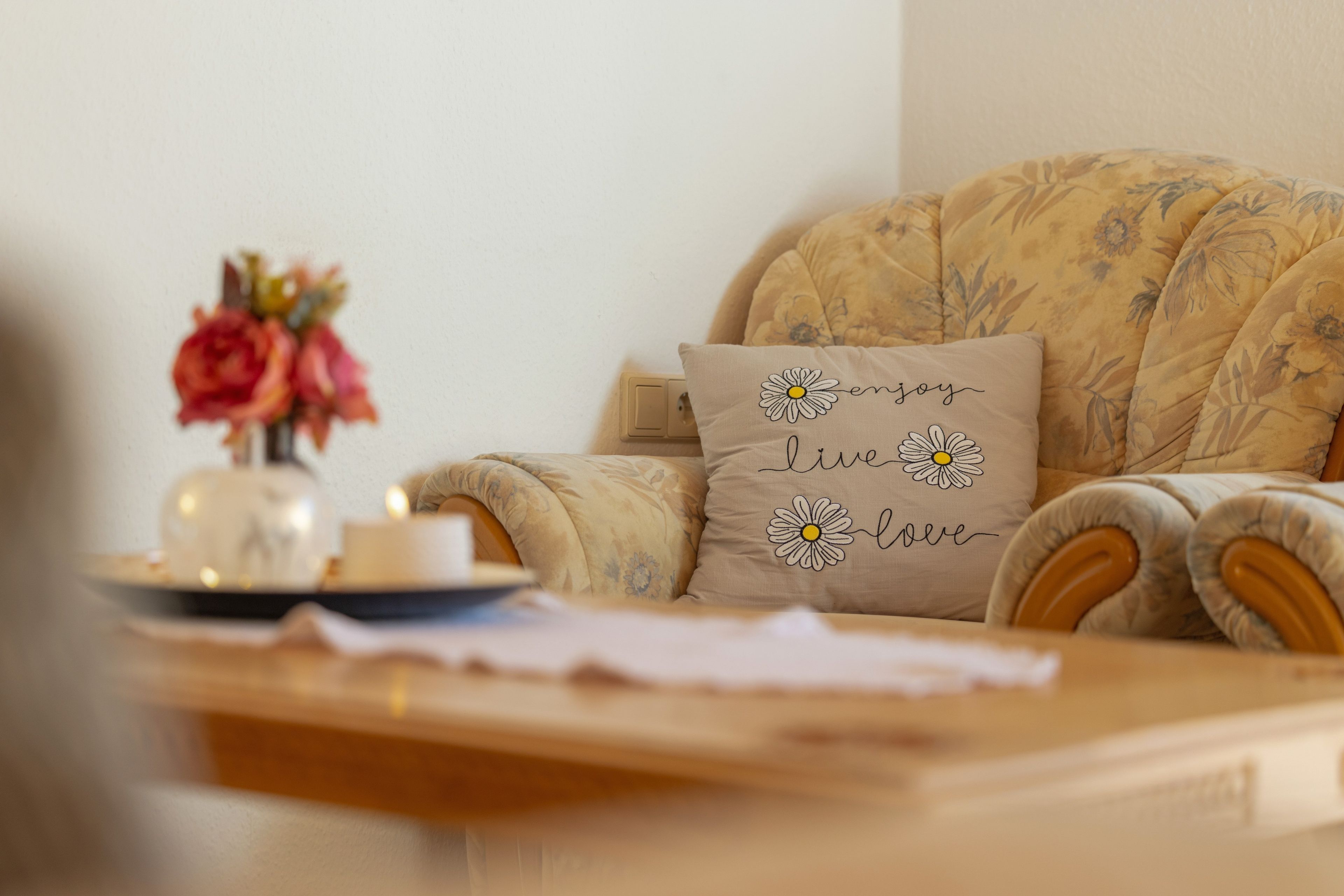 A cozy living room with a floral-patterned sofa, a cushion with a daisy design, and a vase with flowers on a side table.