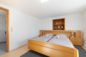 Een slaapkamer met een houten bed, een nachtkastje, een plank met boeken en een deur.