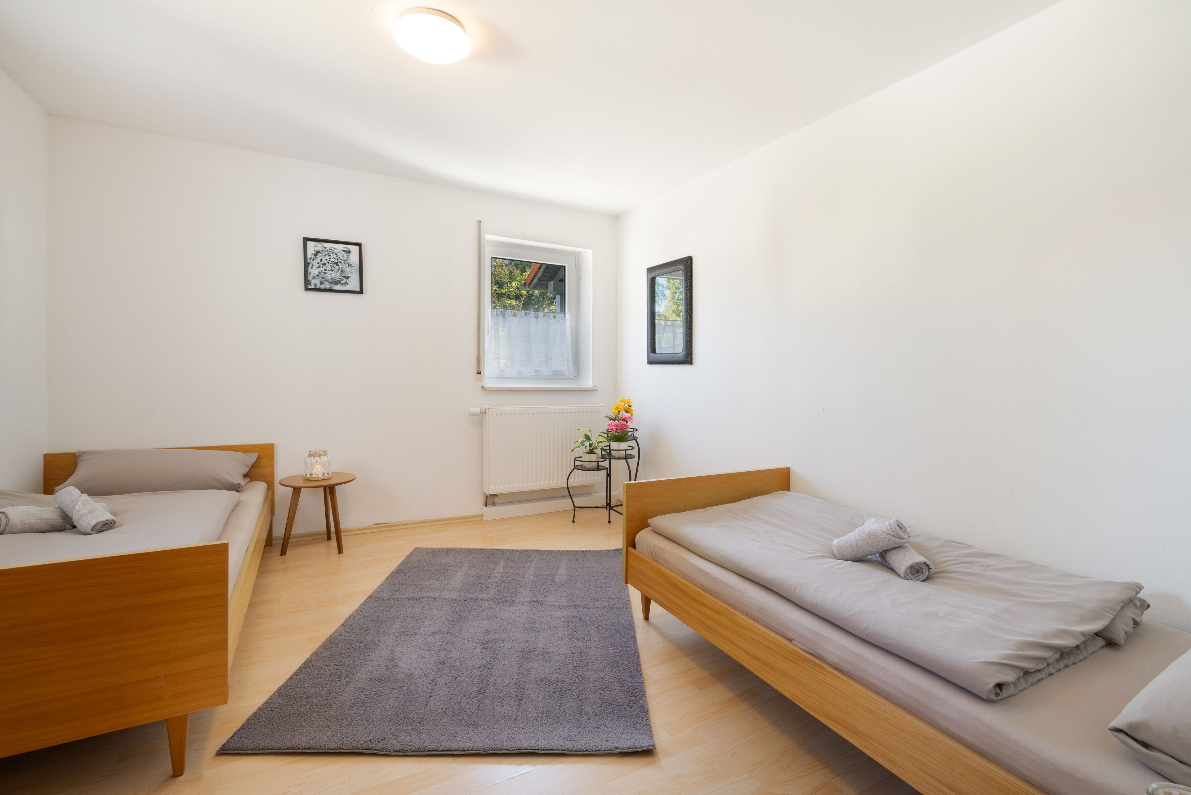 A small, well-lit room with two single beds, a small table with a lamp, a radiator, and a window with a view of trees.