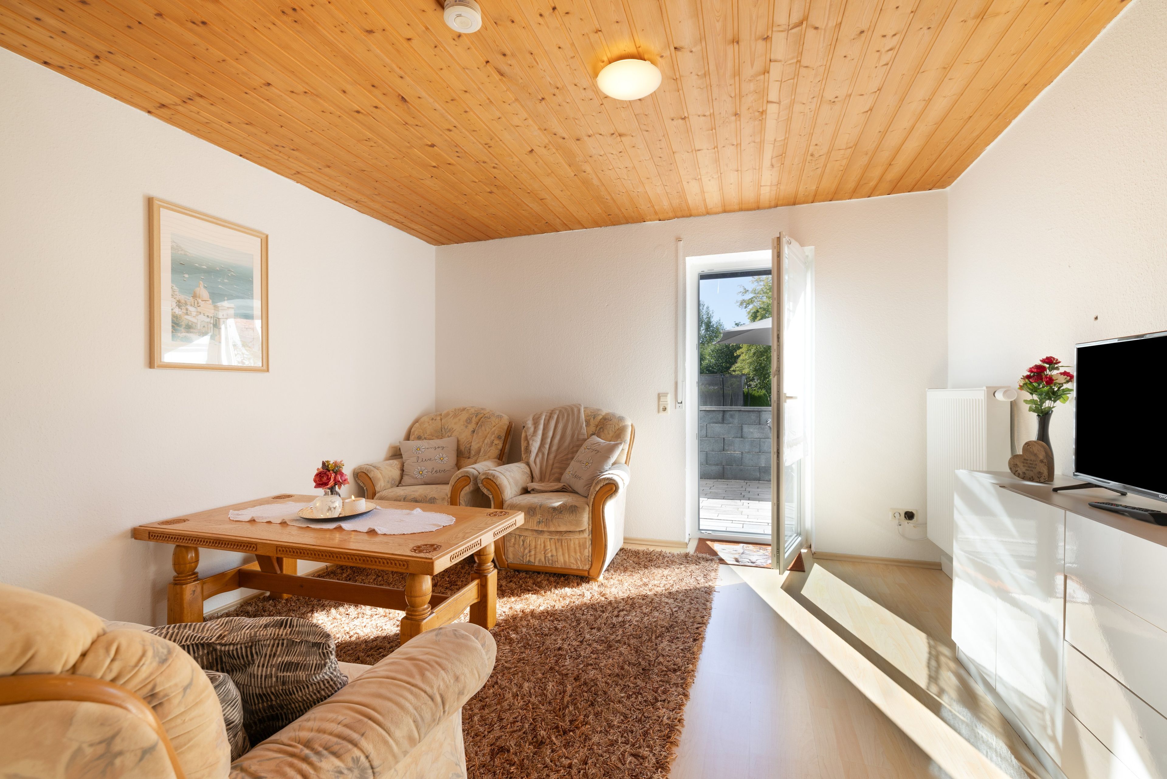 A cozy living room with a wooden ceiling, a dining table with a vase of flowers, two sofas, and a television on a stand.