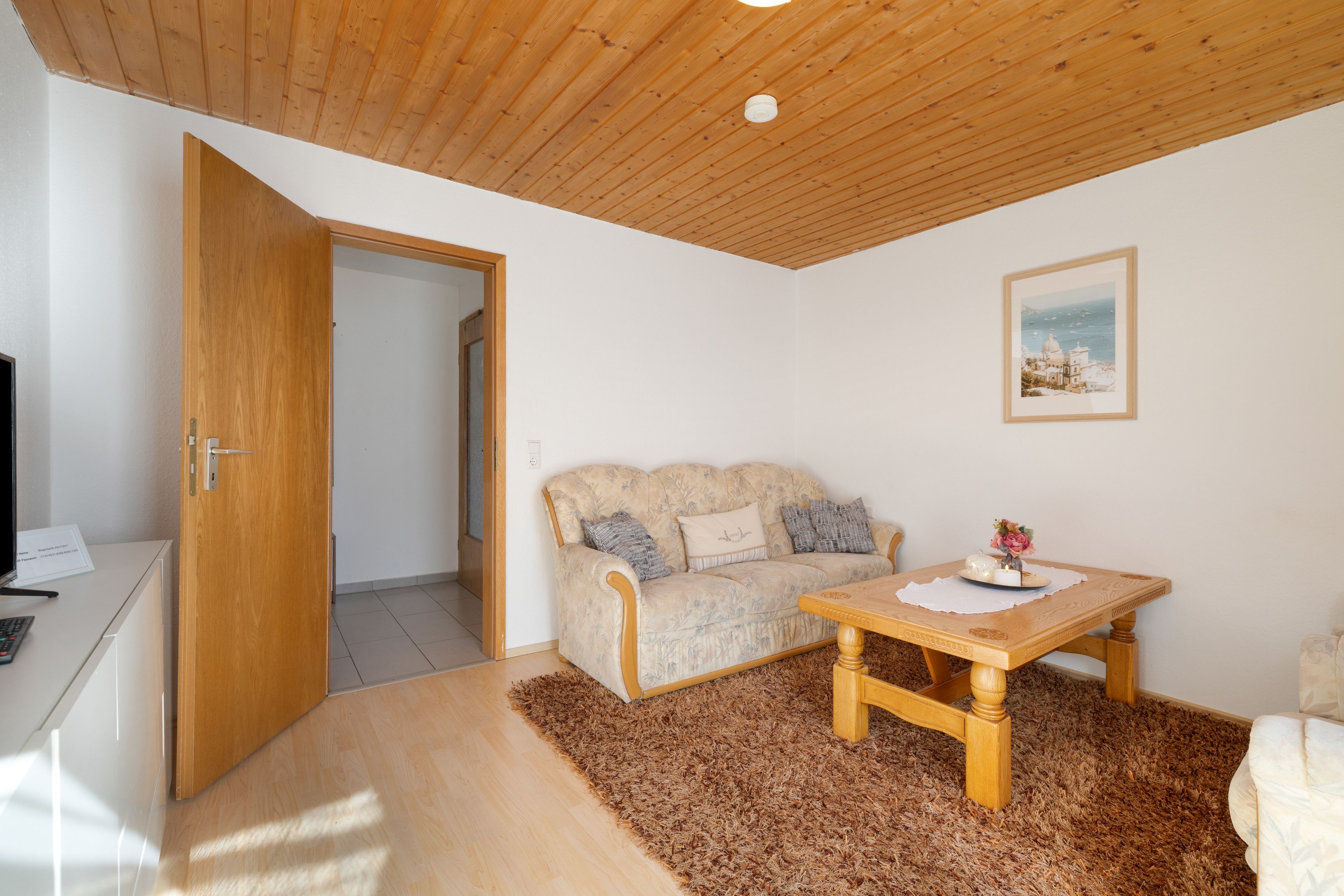 A room with a wooden ceiling, a sofa, a table with a placemat, and a framed picture on the wall.