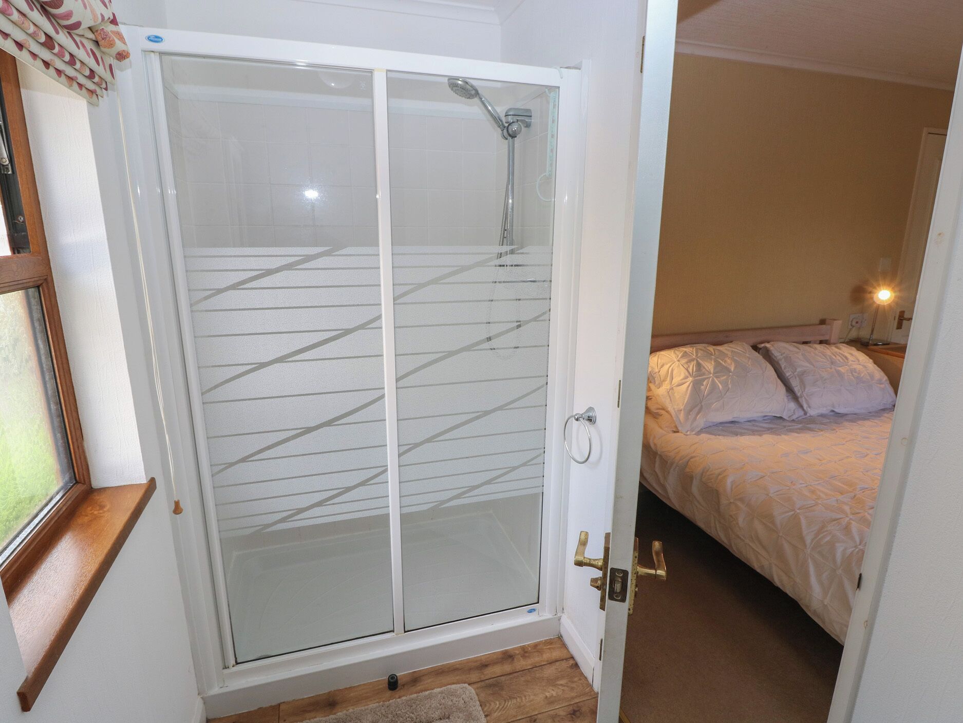 Une chambre dotée d’une porte vitrée donnant sur une zone douche, d’un lit avec une couette blanche et d’une lampe de chevet.
