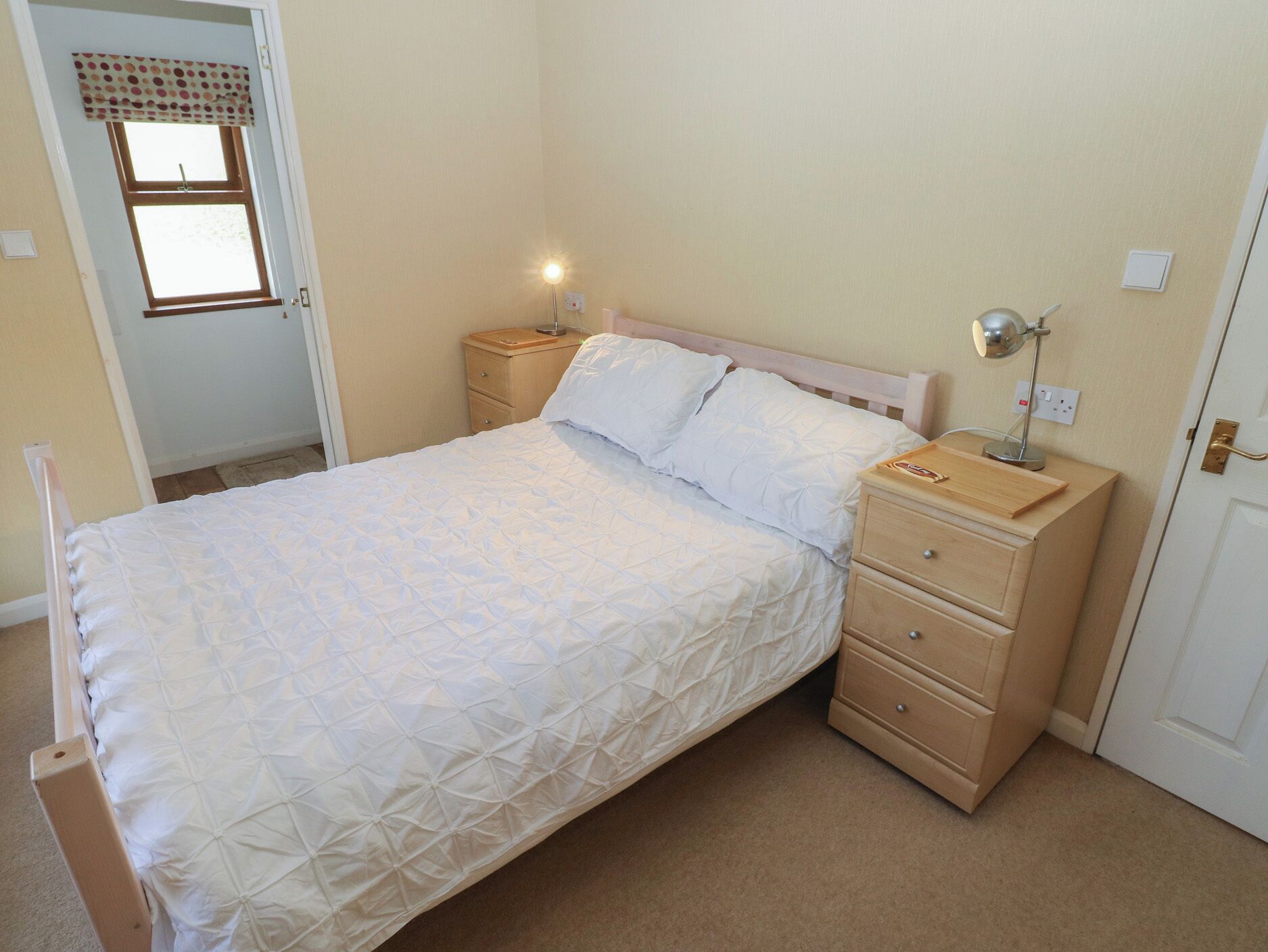 A single bed with a quilted cover, two bedside tables with lamps, a wooden headboard, and a window with a curtain.