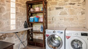 A laundry area with a washing machine, an ironing board, and a wooden shelf with various containers. - Elegant Chania Villa for 4 Guests (Kissamos)